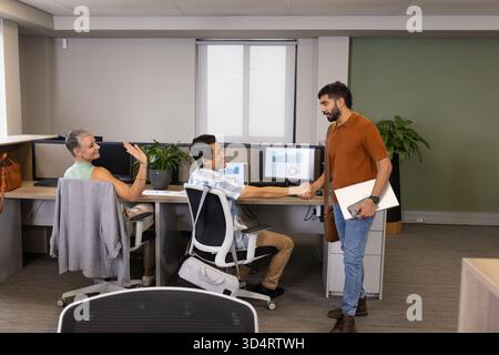 Shaking hands Diverse coworkers standing at desk in office, showing monitors and laptop Stock Photo