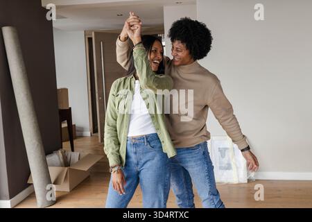 Smiling diverse couple dancing in new home, with cardboard box and wrapped picture frames Stock Photo