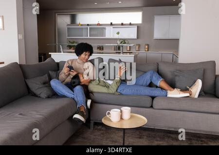 Relaxing Diverse couple using smartphones on gray sectional sofa in living room, with pink mugs Stock Photo
