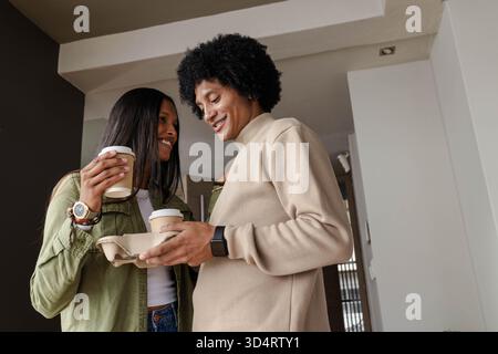 Smiling Diverse couple holding coffee cups in cardboard carrier in home hallway, wearing smartwatch Stock Photo