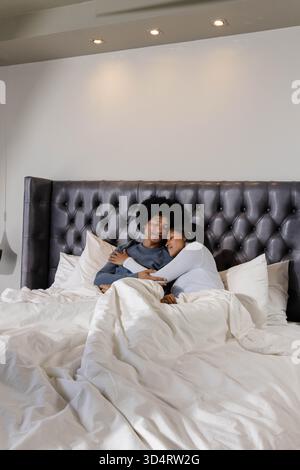 Lying Diverse couple hugging on bed in cozy bedroom, tufted headboard and fluffy pillows Stock Photo