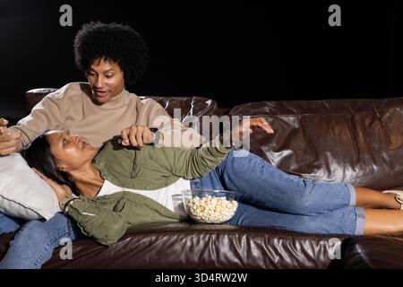 Relaxing Diverse couple cuddling on leather couch on black background, with glass bowl of popcorn Stock Photo