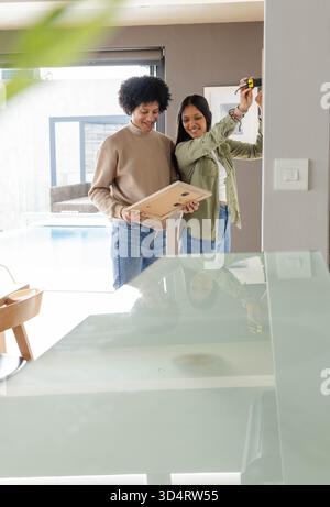 Diverse couple hanging picture frame in modern home interior, using yellow tape measure Stock Photo