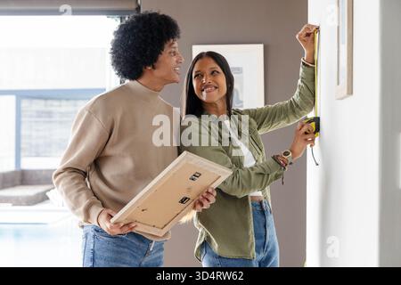 Using yellow tape measure, diverse couple hanging framed picture on neutral wall in living room Stock Photo