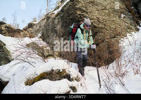 Caucasian young adult man hiking through snowy mountain terrain using trekking poles, wearing backpack and sunglasses, navigating rocky landscape with determination and focus Stock Photo