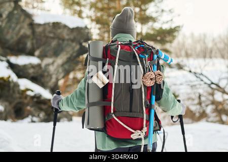 Middle aged Caucasian man hiking in snowy forest carrying large backpack with trekking poles, camping gear and rope visible, standing outdoors facing away from camera in winter landscape Stock Photo