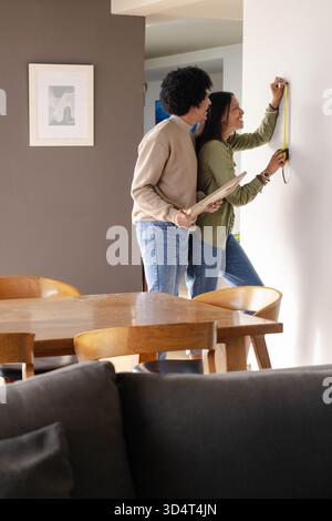 Measuring Diverse couple hanging picture frame on wall in open-plan dining area, with tape measure Stock Photo