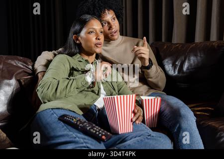 Sitting Diverse couple watching TV on sofa at home, holding striped popcorn buckets and remote Stock Photo