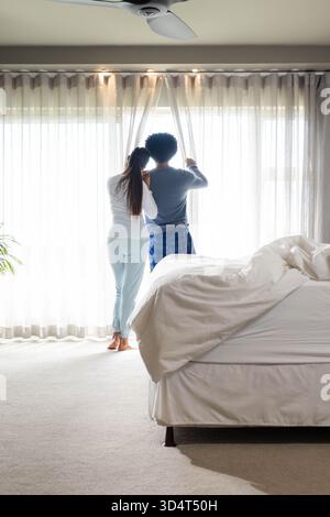 Diverse couple standing pulling sheer curtains in bright bedroom, with unmade bed and potted plant Stock Photo