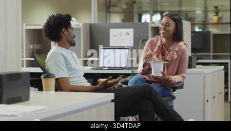 Chatting colleagues at office desks, woman in pink blouse holding takeout using chopsticks, laptop Stock Photo