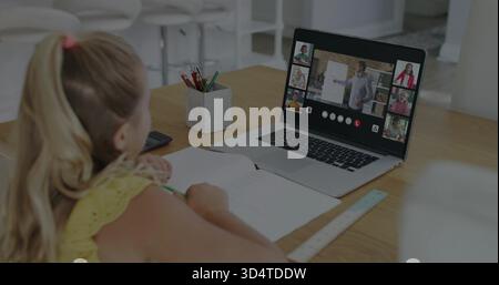 Writing child in yellow top using notebook at home table, laptop showing teacher, copy space Stock Photo