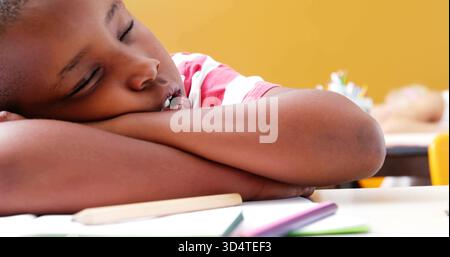 Resting child wearing red-striped shirt leaning on arms at school desk with colored pencils nearby Stock Photo