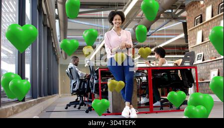 Woman in pink blouse leaning on red desk, open office, holding tablet, green gold heart balloons Stock Photo