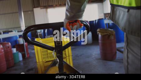 Gripping gloved worker in safety vest using black pallet jack handle in workshop, with yellow crate Stock Photo