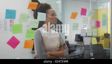 Holding green marker, woman in light blouse studying sticky notes on glass in open-plan office Stock Photo