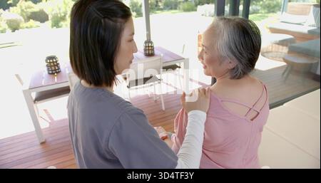 Sitting mother wearing pink strappy top talking with daughter in grey and white on wooden deck Stock Photo