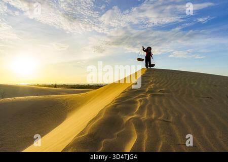 View of a person standing on a golden sand dune under a bright sky with a basket hanging from their shoulder, silhouetted against the setting sun, Tho Stock Photo