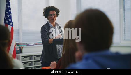 Enthusiastic Smiling Woman Explains Concept Students During Interactive Classroom Discussion. Innovative US Education System, Active Learning, Student Participation, Communication Based Teaching. Stock Photo