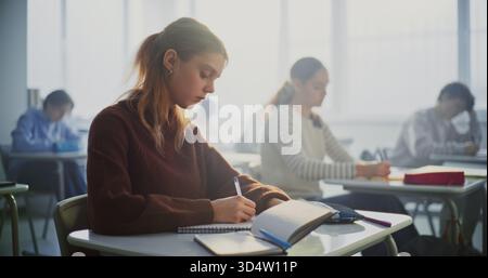 Teenagers Writing Completing Task in Classroom. Diverse Group of Students Sits Desks in Focusing Intently on Writing Assignments. Concept Student Concentration, Academic Workload in Modern Education. Stock Photo