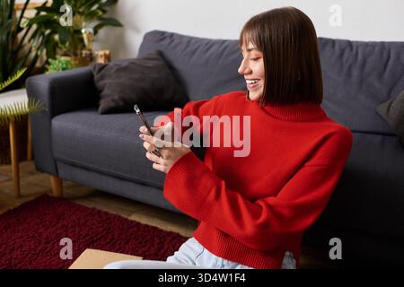 Young woman dressed in a cozy red sweater smiles while using her smartphone at home. Stock Photo