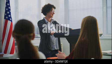 American Teacher Stands Podium Beside US Flag, Engaging Students In Open Discussion About National Values, Civic Responsibility. Concept Patriotic Education, Identity, Positive Learning Environment. Stock Photo
