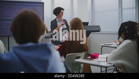 Woman Computer Science Teacher Demonstrates Programming Logic On Big Screen During Classroom Lesson. Concept Coding Education, Digital Literacy, Technology Integration, And Modern Teaching Approach. Stock Photo