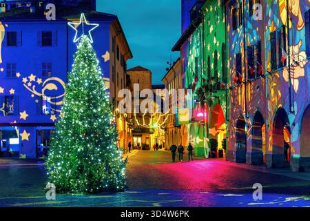 Christmas lights illuminate a festive street with a large decorated tree and colorful projections on historic buildings at night in Alba, Italy. Stock Photo