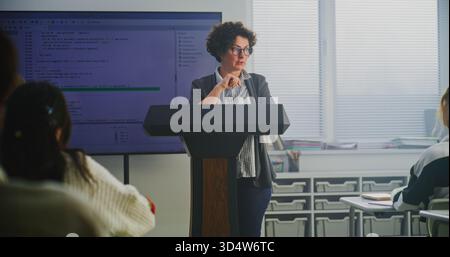 Female Coding Teacher Stands at Podium, Explaining Programming Syntax on Large Digital Screen. Concept Modern Computer Science Education, STEM Learning, Technology Literacy, and Teaching Innovation. Stock Photo