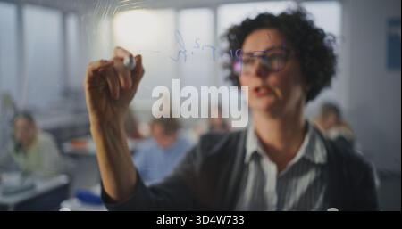 Dynamic Female Teacher With Glasses Writes With Marker Formulas, Addressing Class With Engaging Expression. Concept of Clear Communication, and Teacher Role in Fostering Student Comprehension. Stock Photo