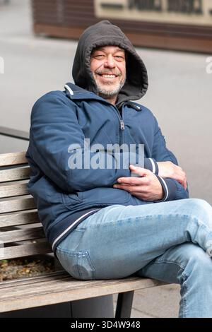 A 50-55-year-old man in a blue jacket and hood is sitting on a bench in the city Stock Photo