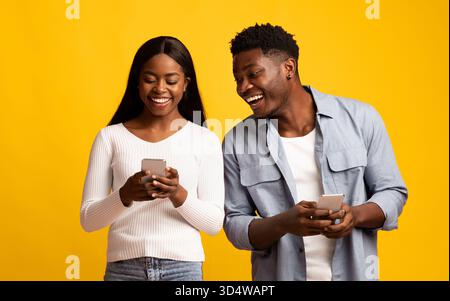 Happy Young Couple Sharing Laughs While Using Smartphones Against a Bright Yellow Background Stock Photo