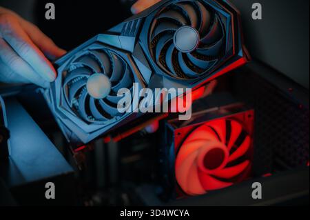 Hands installing graphics card inside desktop, focused DIY technician aligning high-performance GPU above glowing red case fan, close-up low-light sce Stock Photo