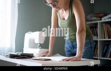 Woman seamstress leaning over her worktable, carefully inspecting the sewing pattern placed on the material in her workshop. Stock Photo