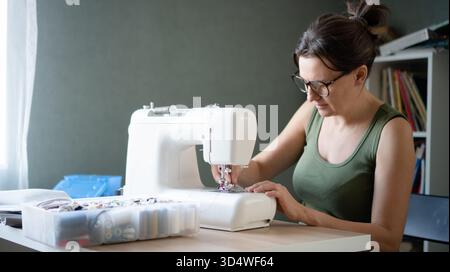 Female tailor using a professional sewing machine to create and assemble a new garment in her bright workshop studio. Stock Photo