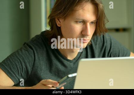A young man with long hair sits at his desk working intently on a laptop. His gaze is focused on the computer monitor. Home office and remote employment concept. Stock Photo