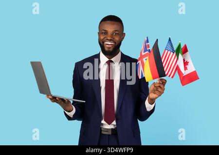 Business Language Courses. Black Businessman Holding Bunch Of Flags And Laptop Stock Photo