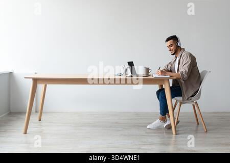 Smiling Arab Man Studies Online While Writing Notes and Using Laptop at Desk Stock Photo