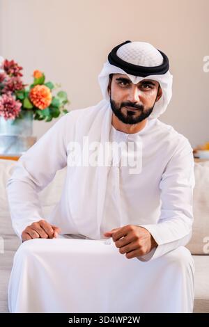 Emirati man with a friendly expression sitting on a sofa in his comfortable home, wearing traditional middle eastern kandura, ghutra, and agal, presen Stock Photo
