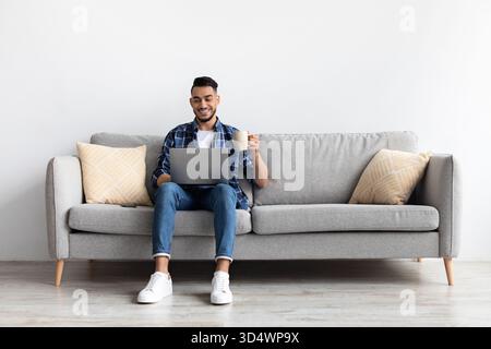 Smiling Arab Man Enjoys Coffee While Watching Video on Laptop From a Comfy Couch Stock Photo