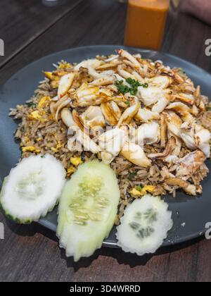Delicious Thai crab fried rice served on a dark plate with fresh cucumber slices on a rustic wooden table. A refreshing iced tea is blurred in the bac Stock Photo