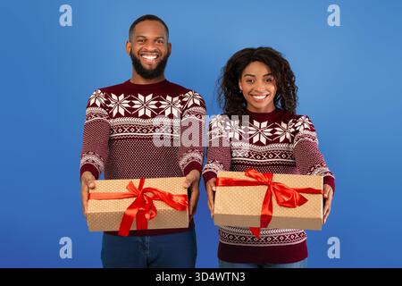 Happy african american couple stretching Christmas gifts to camera over blue studio background, celebrating New Year Stock Photo