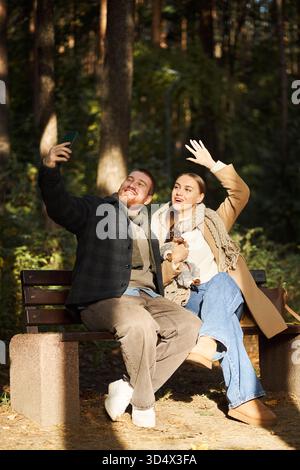 Caucasian young adult man and Caucasian young adult woman sitting on park bench taking selfie with smartphone, smiling and waving, small dog sitting on woman's lap, forest background Stock Photo