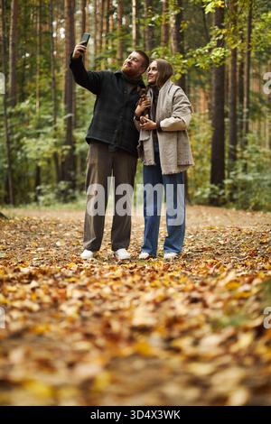 Caucasian young adult man and Caucasian young adult woman standing together in autumn forest taking selfie with smartphone, both looking at device surrounded by fallen leaves Stock Photo