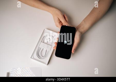 Top view of new iPhone 16 pro resting on white packaging, displaying apple logo on dark screen, accompanying charging cable visible beside device. Stock Photo
