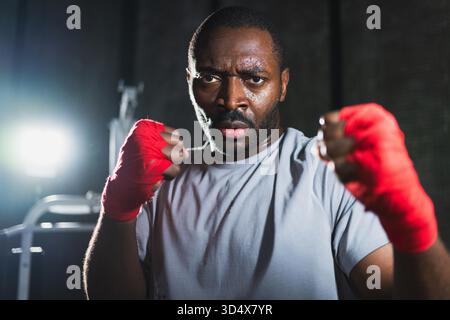 Boxer in gym. Aggressive African man fighter resting after training boxing ready for fight looking at camera. Strong sweated man training punches looking concentrated straight preparing for sparring || Model released Stock Photo