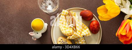 A beautifully arranged Easter table setting showcases a yellow napkin wrapped around a red apple, surrounded by vibrant flowers and a festive bunny decoration, banner Stock Photo