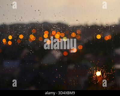 Night-time view of the city seen from inside the window. Raindrops running down the window panes. Vibrant out-of-focus lights in orange and yellow. Stock Photo