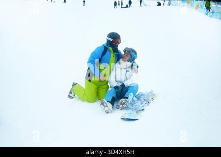 Two people share a moment on the snowy ground at a ski resort, taking a break from snowboarding and enjoying a winter sports day with others around. Stock Photo