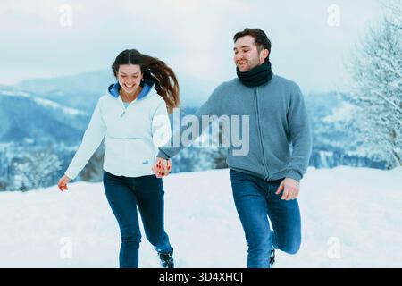 A young couple walks hand in hand through the winter snow, laughing and enjoying the chilly air in a beautiful mountainous landscape. Stock Photo