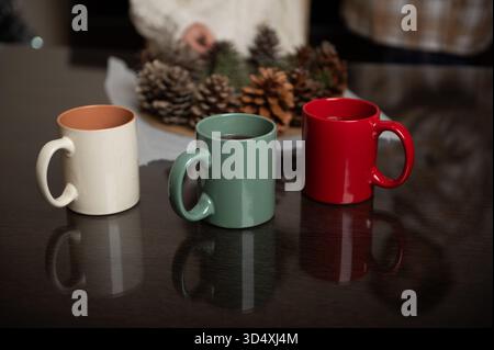 Three ceramic mugs in cream, green, and red sit on a dark wooden table with a pinecone centerpiece in the background, capturing a warm, festive moment Stock Photo